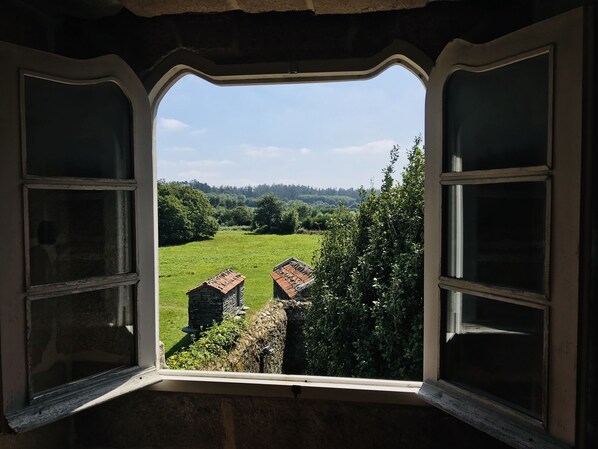 A view through a window with a stone wall and two small buildings in a green field.