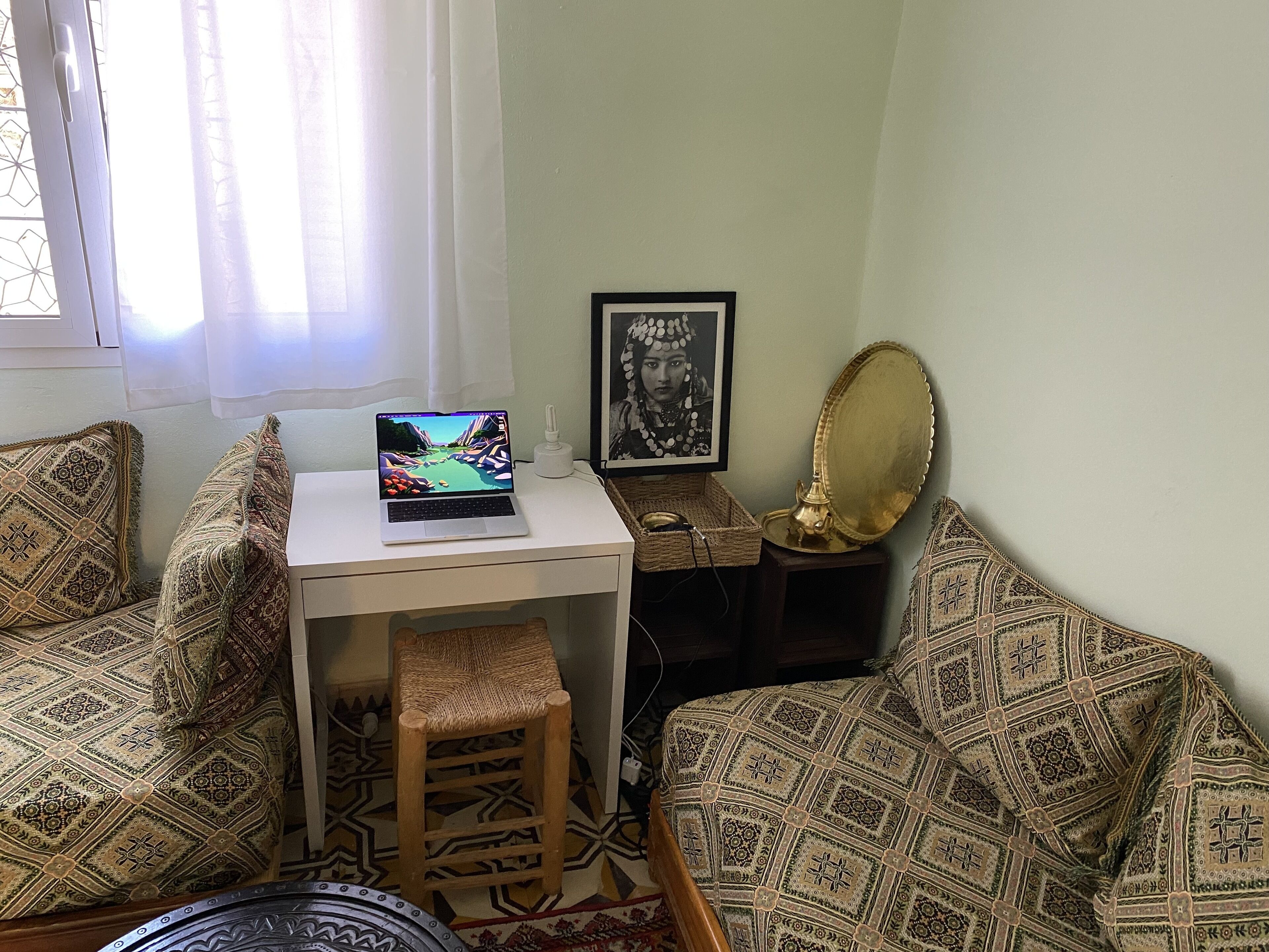 Una habitació petita i acollidora amb un portàtil sobre un escriptori blanc, coixins estampats, una cadira de veta i un mirall decoratiu, a més d'una fotografia emmarcada a la paret.