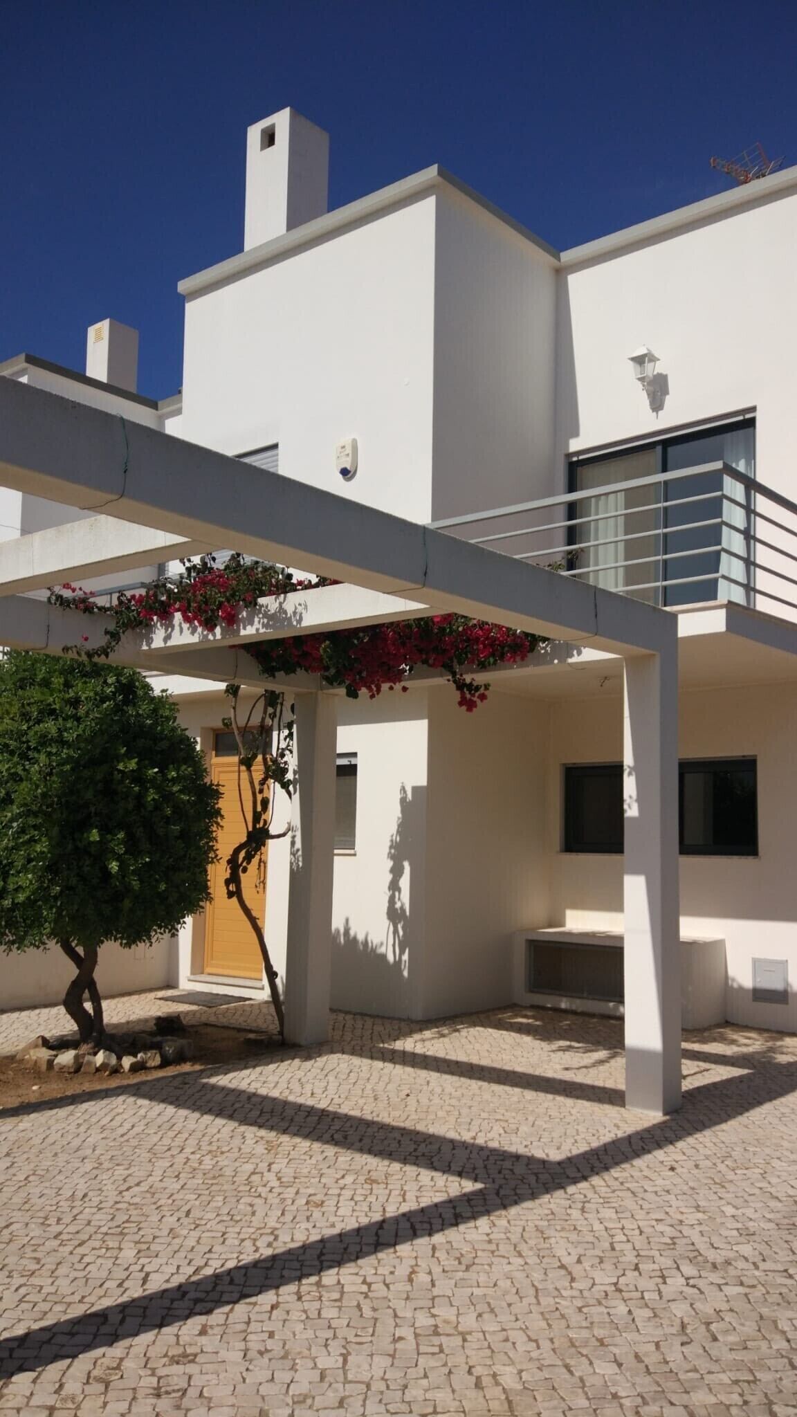 Un edificio moderno con techo plano, un patio cubierto con pérgola y un pequeño árbol.