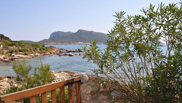 A coastal view with a wooden deck, rocky shoreline, and a mountain in the background. - GARDEN VILLA DOLPHIN SEASIDE (Golfo Aranci)