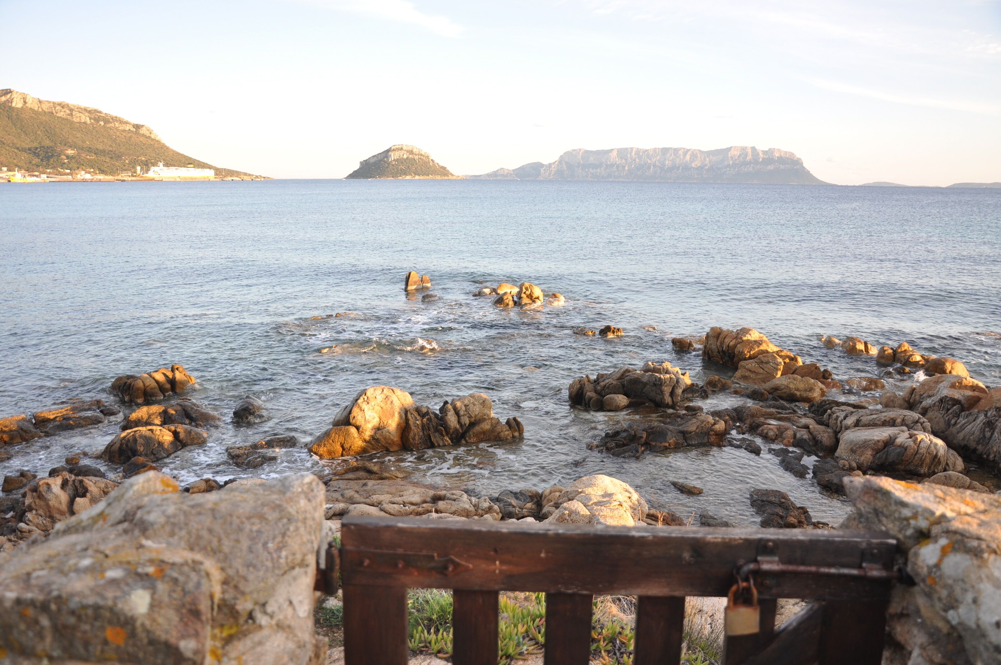 A rocky shoreline with a bench in the foreground, calm water, and distant mountains.