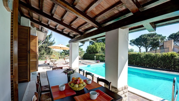 An outdoor dining area with a pool view, wooden table, chairs, and a fruit basket.