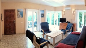 A living room with a sofa, chairs, a table, and a television. There is a door and a window with curtains.
