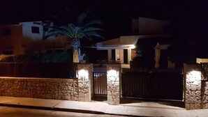 A stone wall with a gate, a palm tree, and a building with illuminated windows at night.