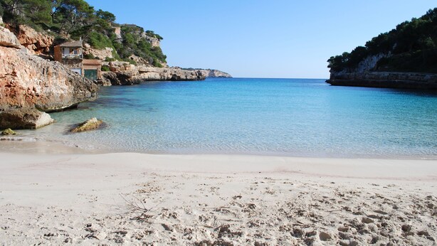 A beach with clear water, rocky cliffs, and a small building on the left.