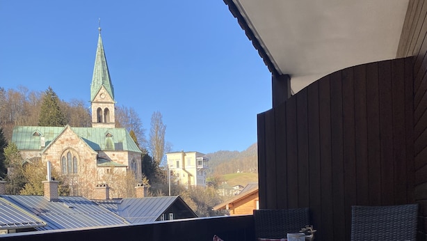 Ein Balkon mit einer hölzernen Bank und zwei Korbstühlen, einem kleinen runden Tisch und karierten Kissen. Im Hintergrund ist eine Kirche mit einem grünen Dach zu sehen.