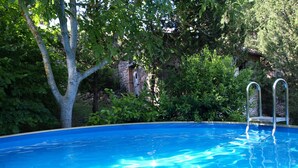 A clear blue swimming pool with a ladder, surrounded by lush greenery and a stone building in the background.