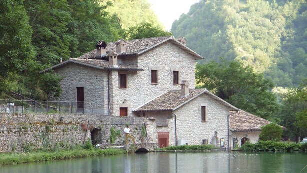 A stone building with a tiled roof, a small bridge, and a body of water in the foreground.