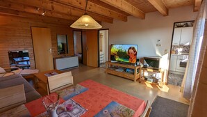 Ein gemütliches Wohnzimmer mit Holzbalkendecke, einem Fernseher, einer Küchenzeile und einem Esstisch mit Unterlage.