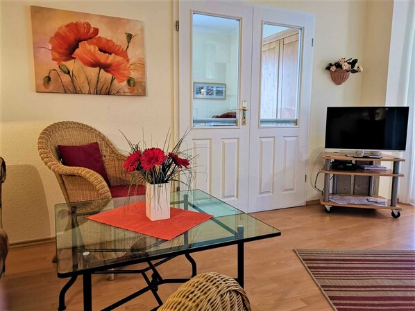 Un salon avec une table à plateau en verre, des fauteuils en osier, un téléviseur posé sur un meuble et un tableau représentant des fleurs.