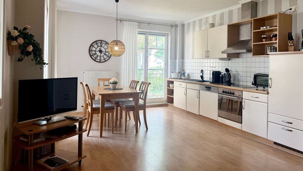 Una cocina moderna con armarios blancos, un comedor con mesa y sillas de madera, y un televisor sobre un mueble.