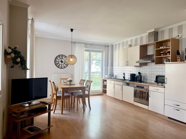 A modern kitchen with white cabinets, a dining area with a wooden table and chairs, and a television on a stand.