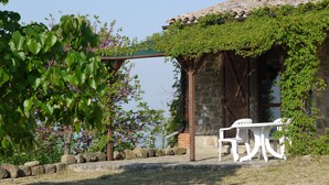 A rustic outdoor seating area with a stone wall, a pergola covered in greenery, and a white plastic chair.