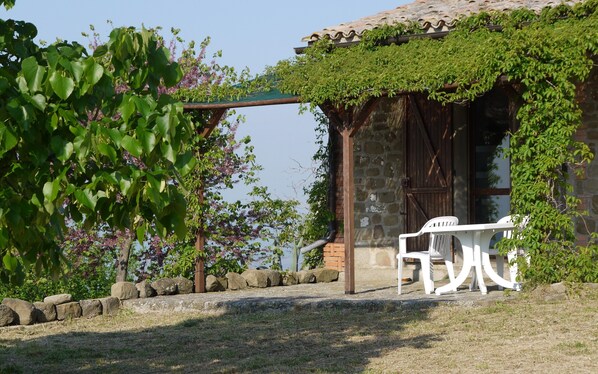 A rustic outdoor seating area with a stone wall, a pergola covered in greenery, and a white plastic chair.
