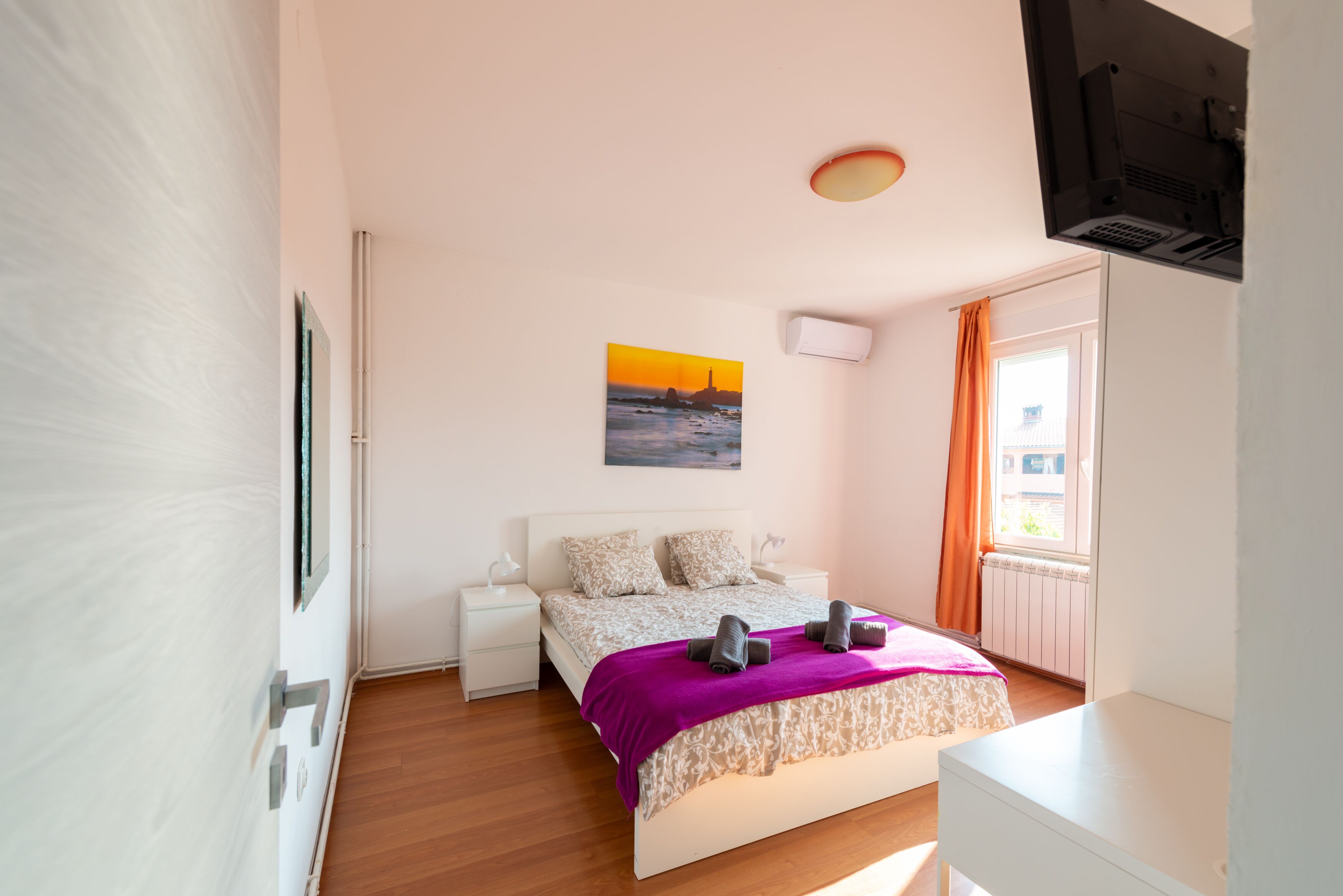 Una camera da letto con un letto, un comodino, una TV a parete, una finestra con tende e un quadro appeso al muro.