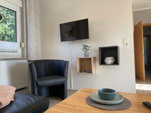 A modern living room with a black leather chair, a wooden table with a bowl, a flat-screen TV mounted on the wall, and a small shelf with a plant and decorative item. - Holiday house Geers am Klostersee Dargun - Holiday home Geers (Dargun)