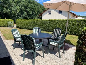 An outdoor patio with a round table and four chairs, surrounded by a hedge and a house in the background.