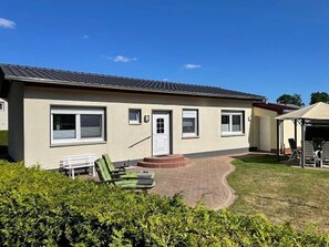 Een laag gebouw met plat dak, een voordeur en ramen. Er is een verhard gebied met een bank en een overdekte zitplaats met parasols.