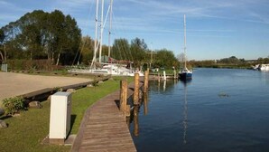 Eine ruhige Marina mit mehreren vertäuten Segelbooten, einem Holzsteg, der zum Wasser führt, und einer grünen Fläche mit Bäumen im Hintergrund.