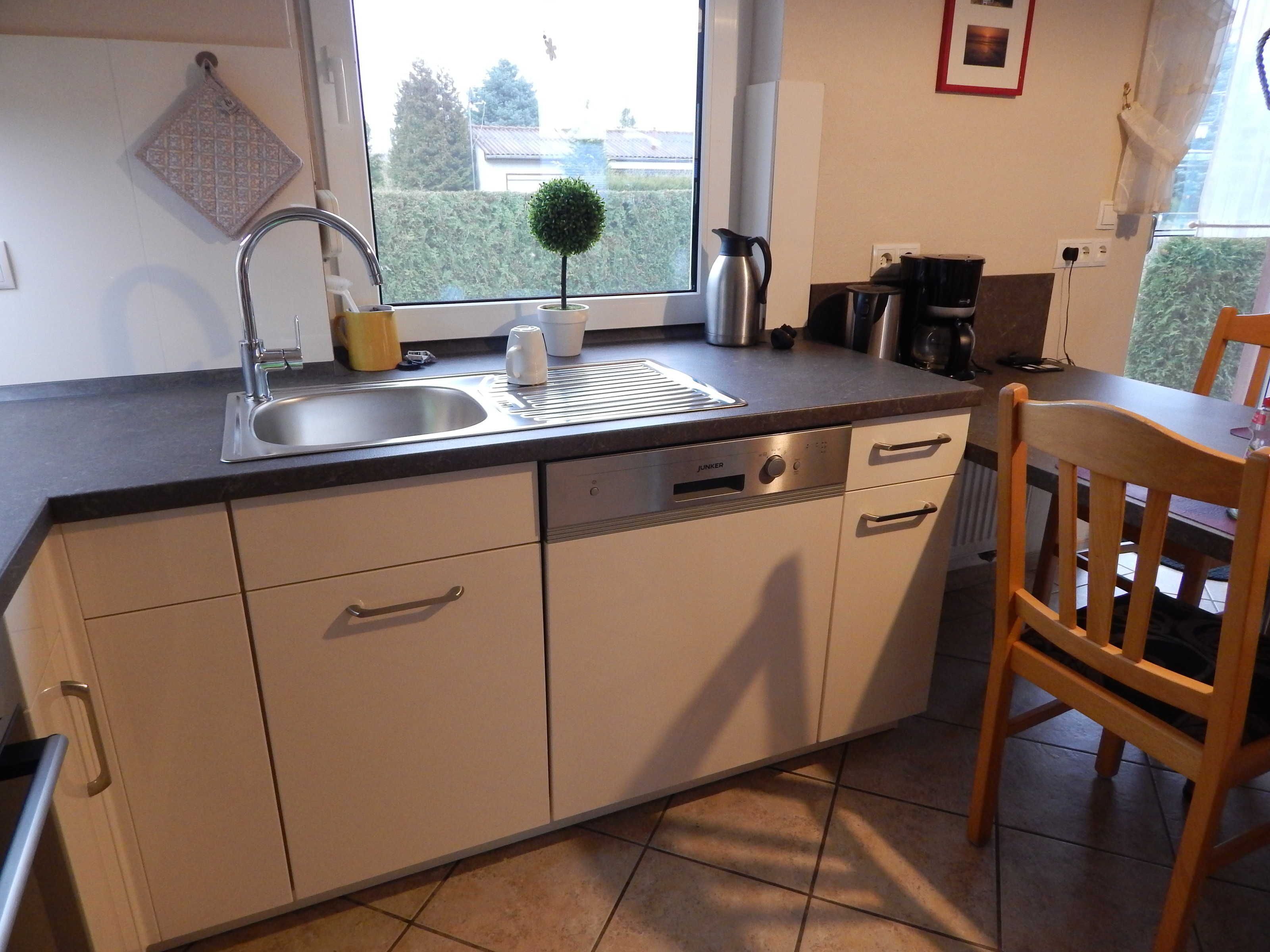 A kitchen with a sink, a small potted plant, a kettle, and a window with a view of greenery outside.
