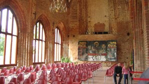 Ein großer Saal mit Reihen roter Stühle, hohen, gewölbten Backsteinwänden und großen Fenstern.
