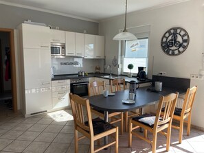 A modern kitchen with white cabinets, a built-in oven, a microwave, and a coffee maker. There is a dining table with four chairs and a clock on the wall.