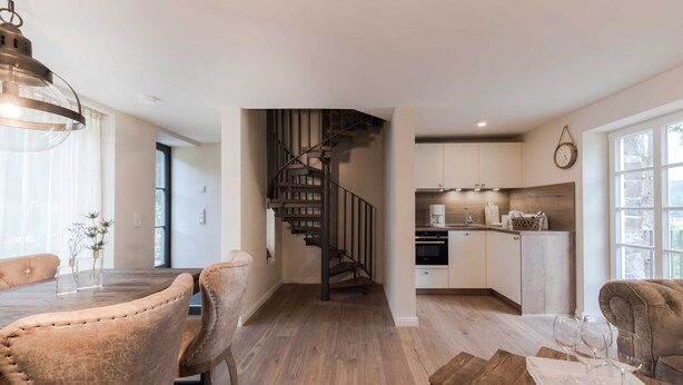 Een moderne keuken met een inbouwoven, een eethoek met een houten tafel en stoelen, en een trap naar een verdieping erboven.