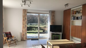 A living room with a sofa, a television on a stand, a bookshelf, and a view of a garden through a sliding glass door. - Modern, quiet apartment in the countryside - in 30 minutes by train to Messe Frankfurt (Friedrichsdorf)