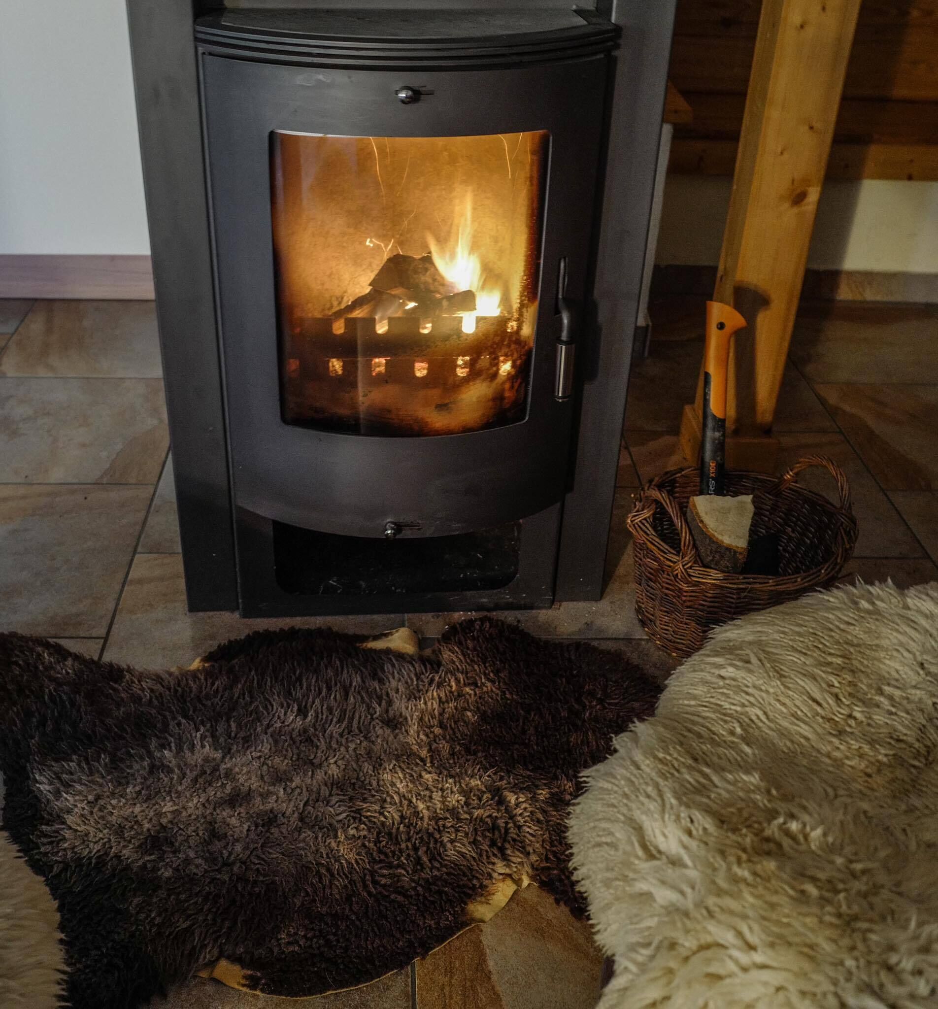 En vedeldad kamin med en glaslucka, en korg och en mjuk, lurvig matta på golvet.