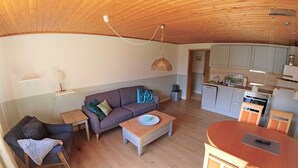 A cozy living room with a sofa, coffee table, and dining area. A kitchen with cabinets and appliances is visible in the background. - Apartment "Schwansen" - Apartments on the holiday farm Jochimsen (Kappeln)