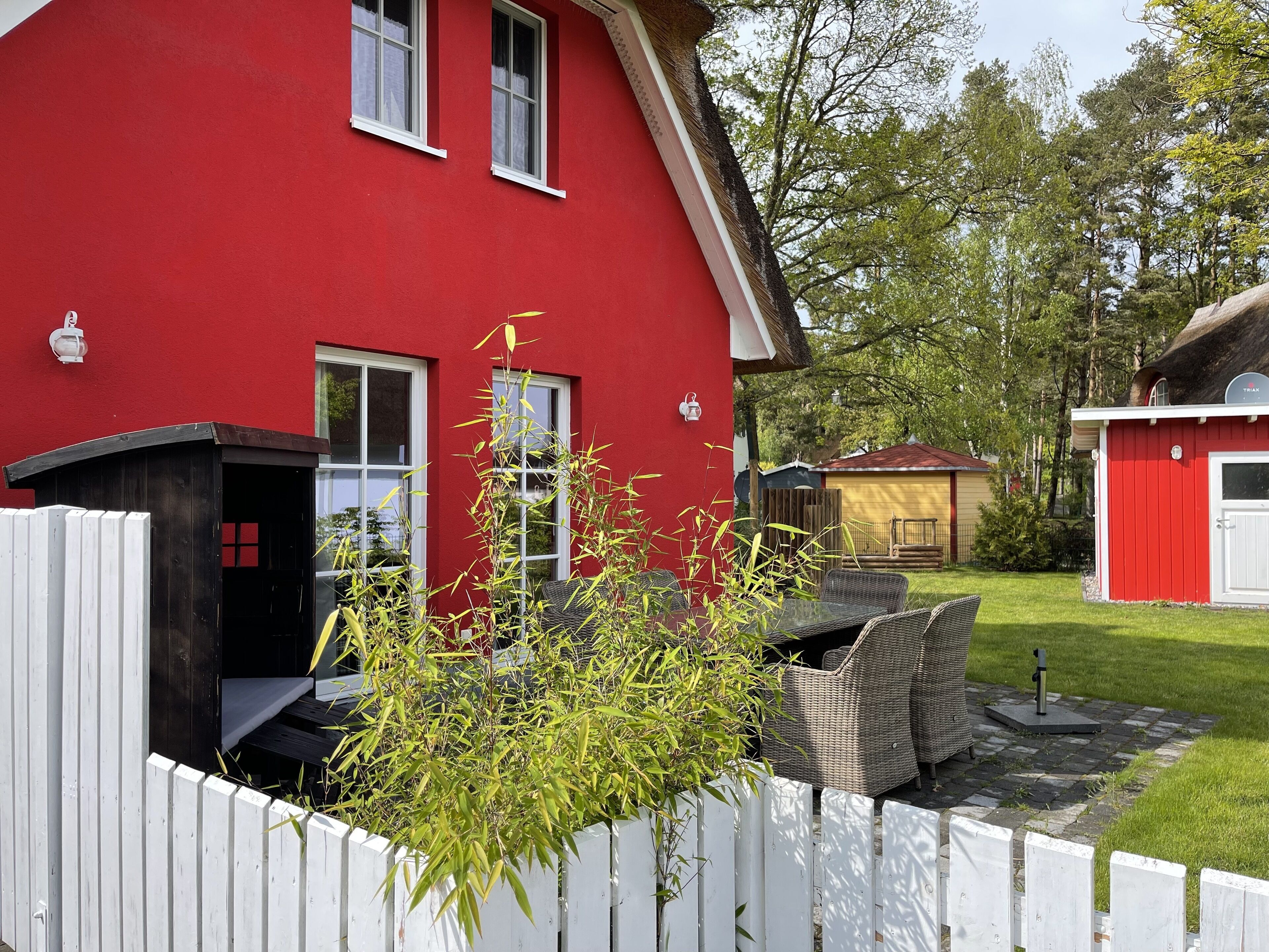A red house with a white fence, a patio with wicker furniture, and a small shed in the background.