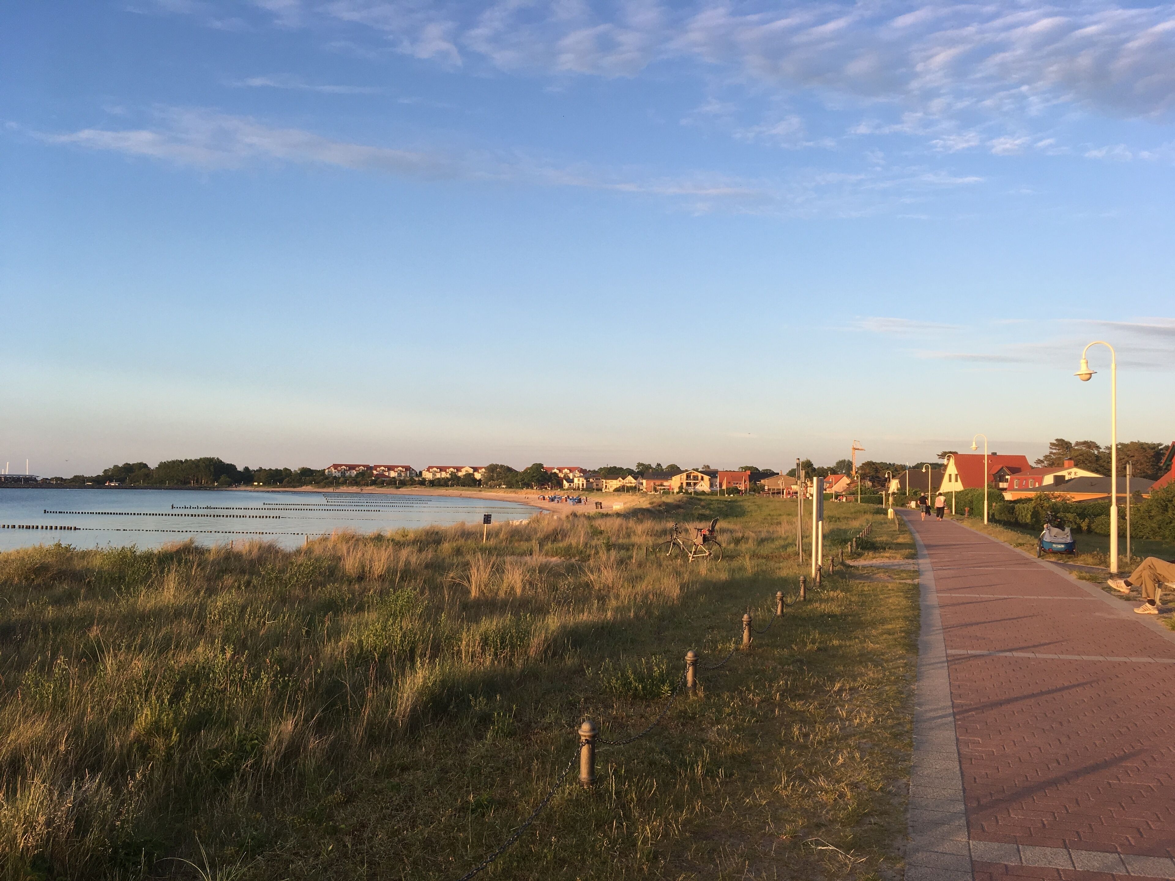 Uma passarela costeira com um caminho de tijolos vermelhos, áreas ajardinadas e casas ao fundo.