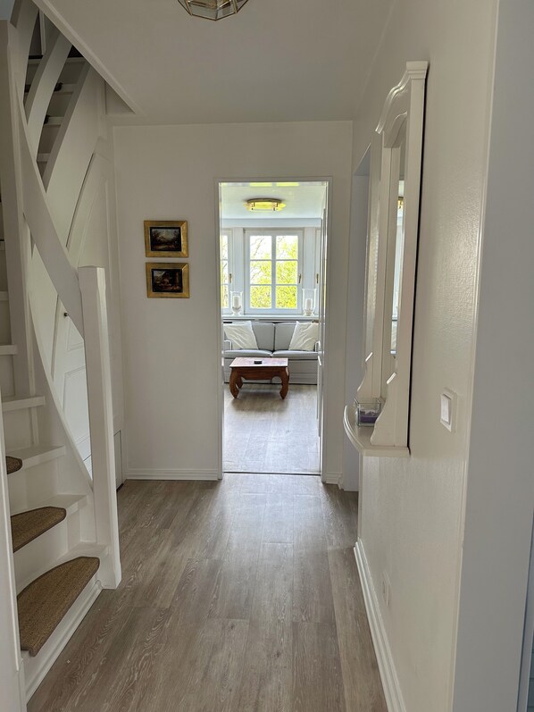 A hallway with a wooden floor, white walls, and a staircase on the left. There is a window with a view of trees and a bench with cushions.
