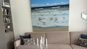 Un salon avec un canapé beige, une table basse en verre surmontée d’un chandelier, une bibliothèque et un grand tableau représentant une plage.
