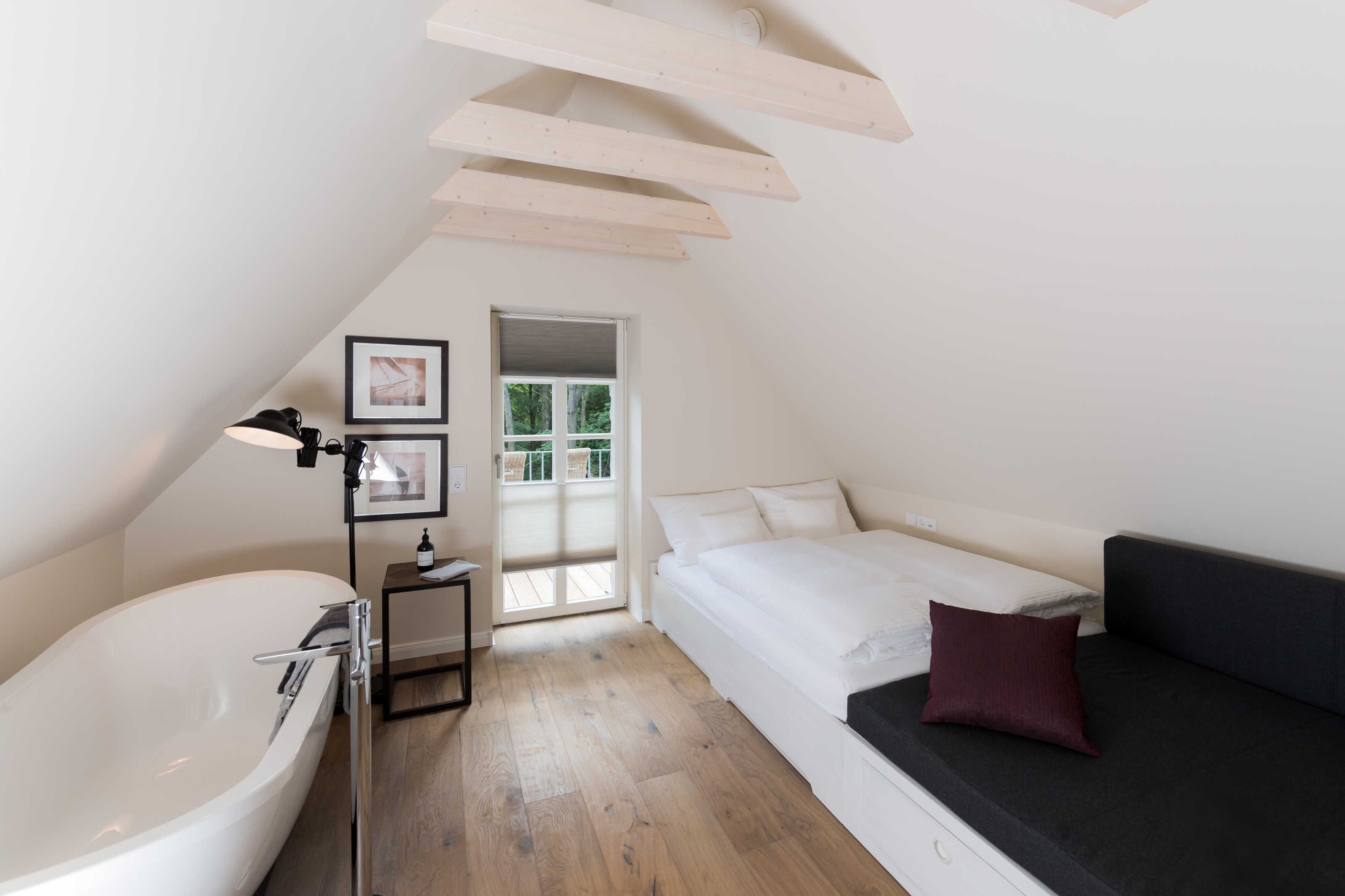 Een slaapkamer met een groot, wit bad, een bed met een donkere hoofdeinde en een klein nachtkastje met een lamp.