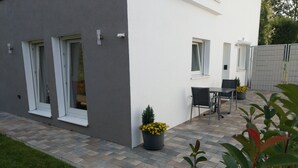 Eine moderne Terrassengrundstück mit Tisch und Stühlen, Pflanzkübeln und einem Weg zu einem Gebäude mit großen Fenstern.