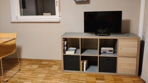 Ein Zimmer mit Fernseher auf einer Holzkassette, einem Fenster und einer Pflanze auf einem Regal.