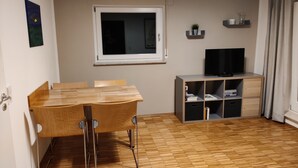 Ein kleiner, gut beleuchteter Raum mit einem Holztisch und Stühlen, einem Fernseher auf einem Regal und einem Fenster mit Blick nach draußen.