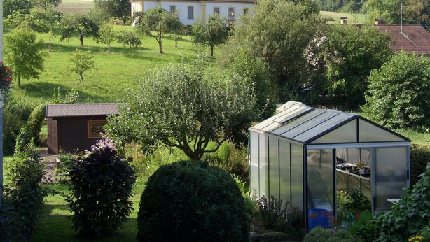 Una casa grande con una torre, rodeada de abundante vegetación y un invernadero.