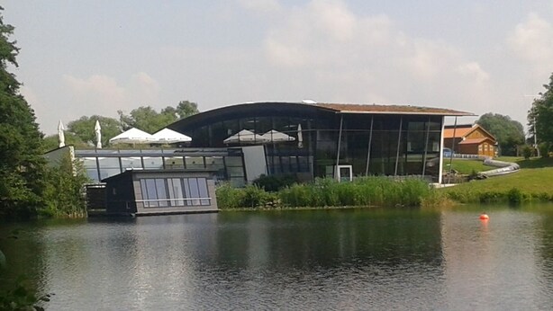 Un edificio moderno con fachada de vidrio, un área cubierta con toldos blancos y un pequeño barco amarrado cerca del agua.