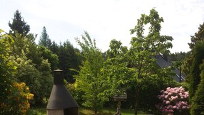 Una parrilla de piedra en un jardín con terraza de madera, rodeada de árboles y plantas con flores.
