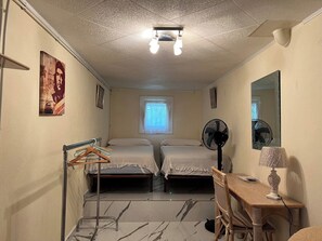 Ein kleiner, kompakter Raum mit zwei Betten, einem Schreibtisch, einem Stuhl, einem Spiegel, einem Ventilator und einem Fenster.