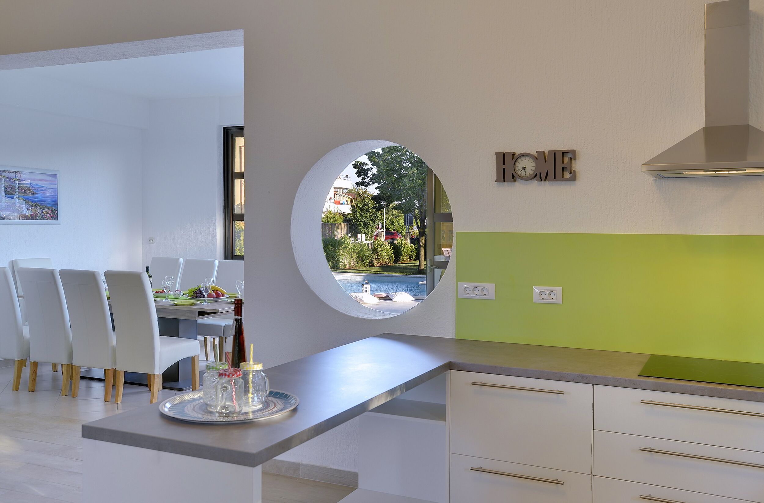 A modern kitchen with a view of a swimming pool, a dining area with white chairs, and a green accent wall.