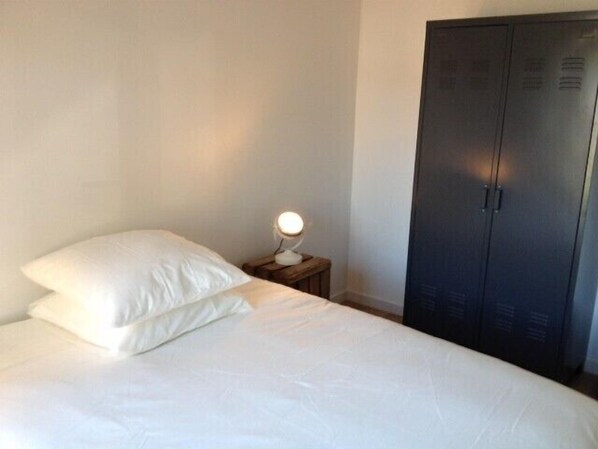 Una cama bien tendida con ropa de cama blanca en una habitación con un armario oscuro y una mesita de noche pequeña con una lámpara.