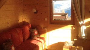 Una cabaña acogedora con interior de madera, un sofá, una mesa y un ventanal con vistas a un paisaje nevado.