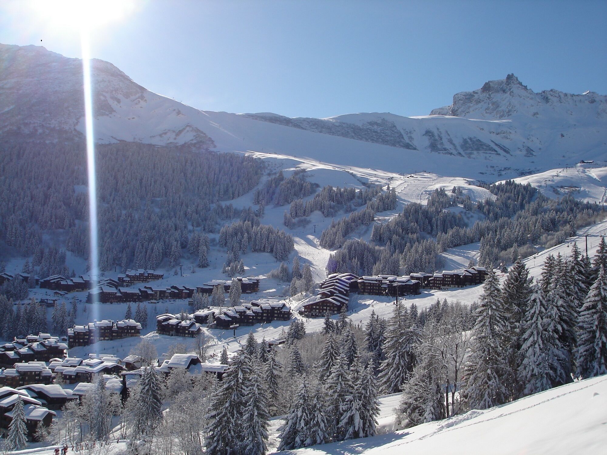 A snow-covered mountain resort with multiple chalet-style buildings clustered together, surrounded by dense forests and steep slopes.