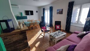 Ein Wohnzimmer mit Holzboden, einer Essecke mit Tisch und Stühlen, einem an der Wand befestigten Fernseher und einem Fenster mit Vorhängen.
