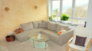 A living room with a sectional sofa, a glass coffee table, a wicker basket with bottles, and a view of trees through large windows. - Exclusive Holiday Apartment with Moselle View & Bike Garage (Bullay)