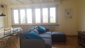 A living room with a grey sectional sofa, a small table with a white tablecloth, and a radiator below a large window. - Aviva (95 m2 apartment with terrace) (Huningue)