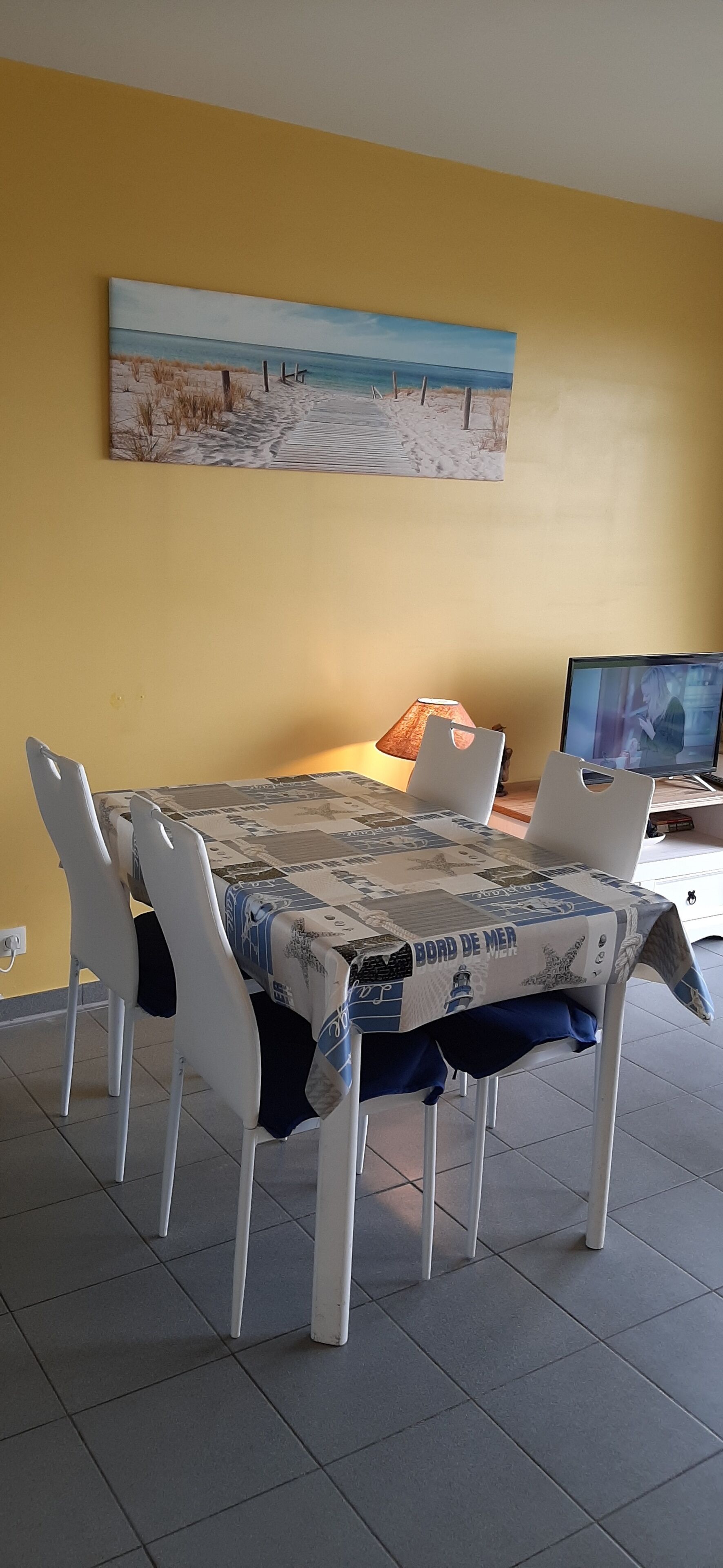 A dining area with a table covered by a nautical-themed tablecloth, white chairs, and a painting of a beach scene on the wall.